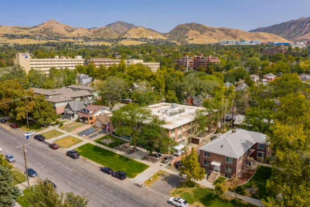 Aerial view of a residential neighborhood with multiple houses and apartment buildings surrounded by trees, with mountains in the background under a clear sky.