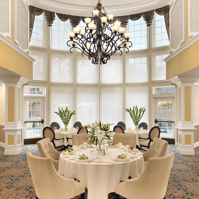 Elegant dining room with a round table set for a meal beneath a large chandelier in front of tall, arched windows.