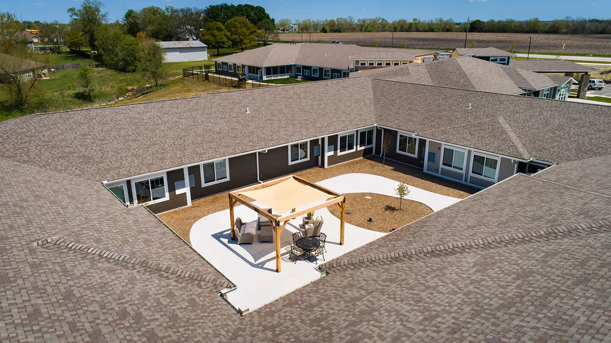 Aerial view of a senior living facility courtyard with a shaded seating area including chairs and a table, surrounded by a single-story building with multiple windows and doors. The courtyard has a paved walkway and some small trees planted in mulch beds. The surrounding area includes open fields and some trees.