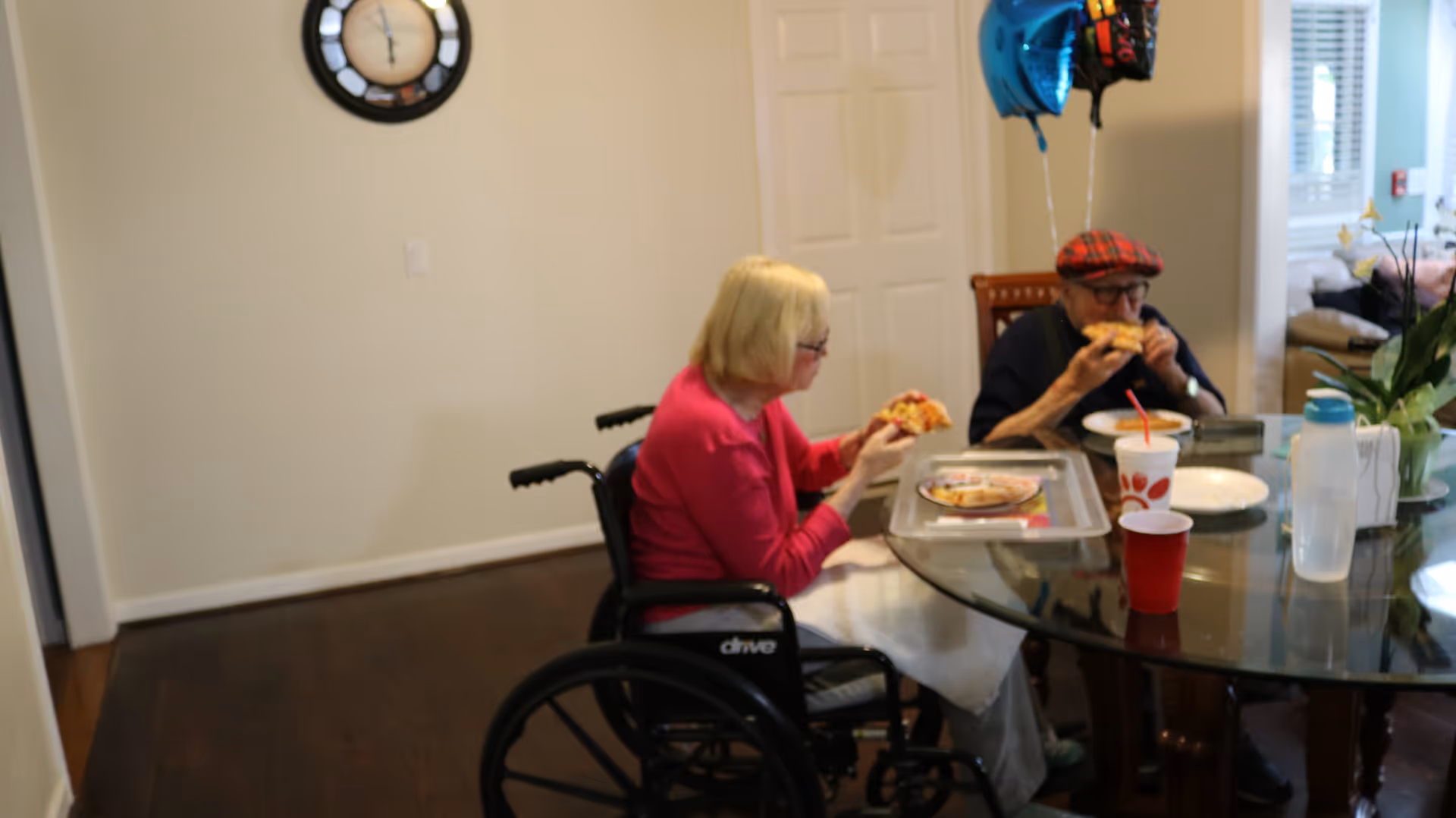 Two elderly individuals sitting at a glass dining table eating pizza. One person is in a wheelchair wearing a pink sweater, and the other is wearing a red plaid hat and glasses. There are balloons tied to the chair, a clock on the wall, and various items on the table including a drink cup with a straw and a water bottle.