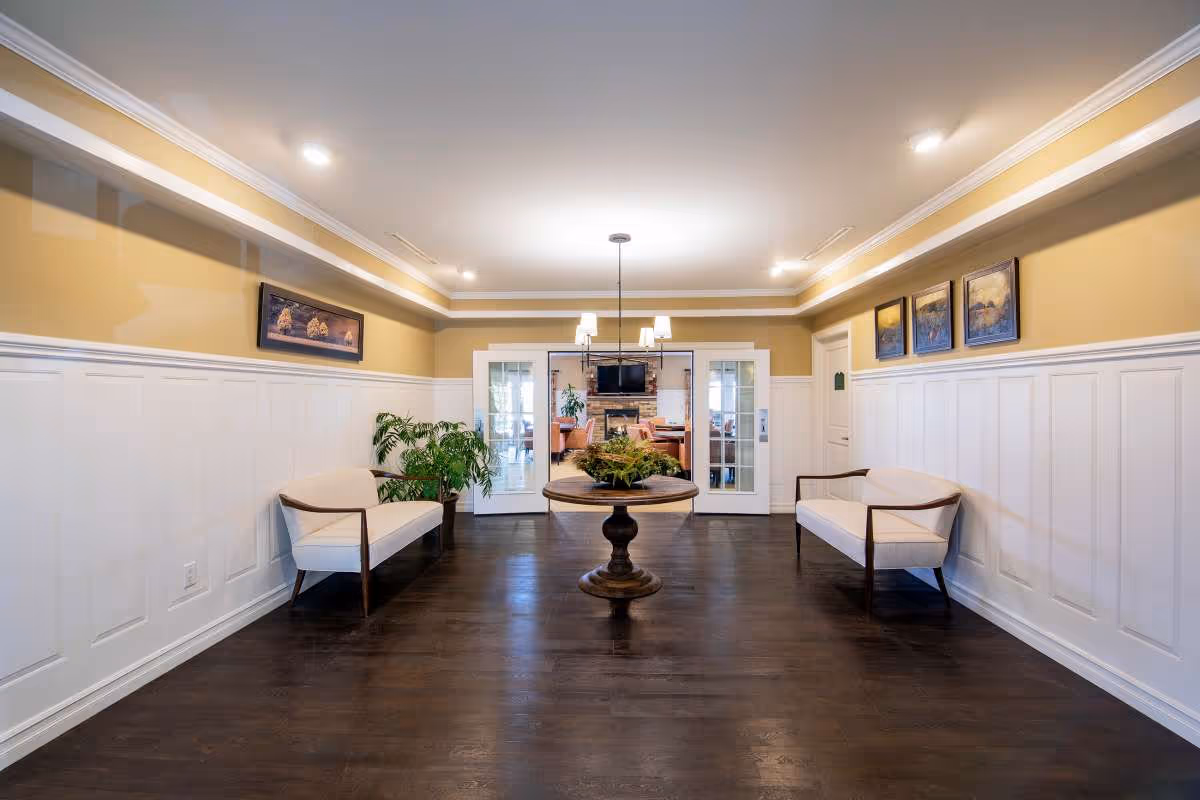 A spacious, well-lit interior hallway with dark wooden floors, cream-colored walls with white wainscoting, and a round wooden table with a plant centerpiece in the middle. Two white cushioned benches with wooden armrests are placed symmetrically on either side of the hallway. The hallway leads to a room with glass double doors, where a fireplace, TV, and seating area are visible. Several framed paintings hang on the walls.