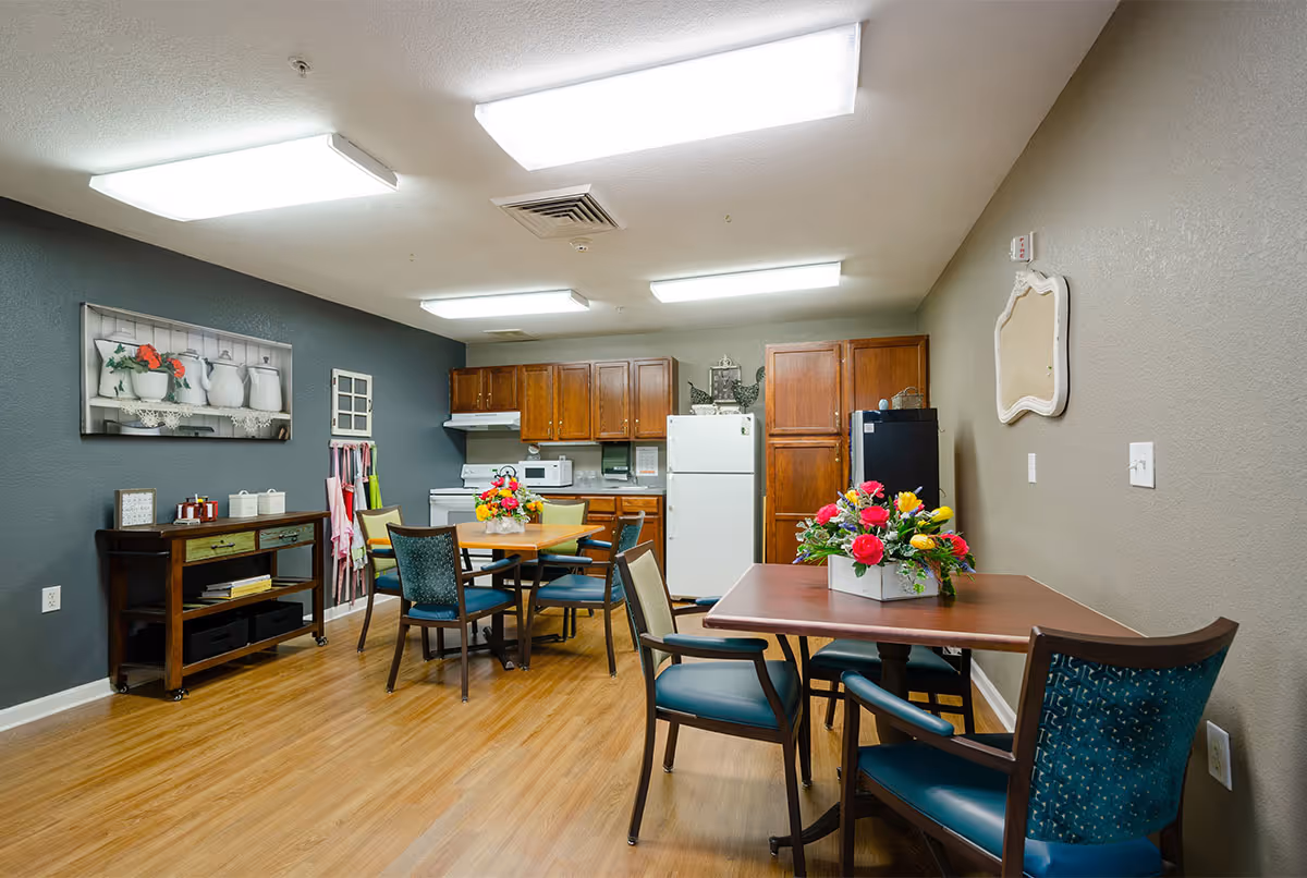 Interior view of a communal kitchen and dining area with wooden cabinets, a white refrigerator, microwave, stove, and two tables with chairs. Each table has a colorful flower arrangement. The room has wood flooring, gray and beige walls, and bright overhead lighting.