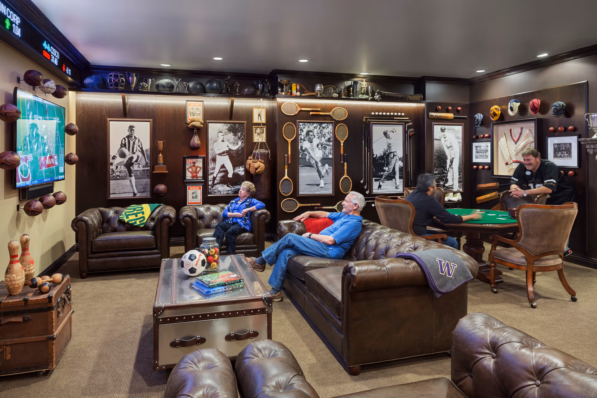 Residents relax in a cozy game room with leather sofas, a wall-mounted TV, sports memorabilia on the walls, and a card table.