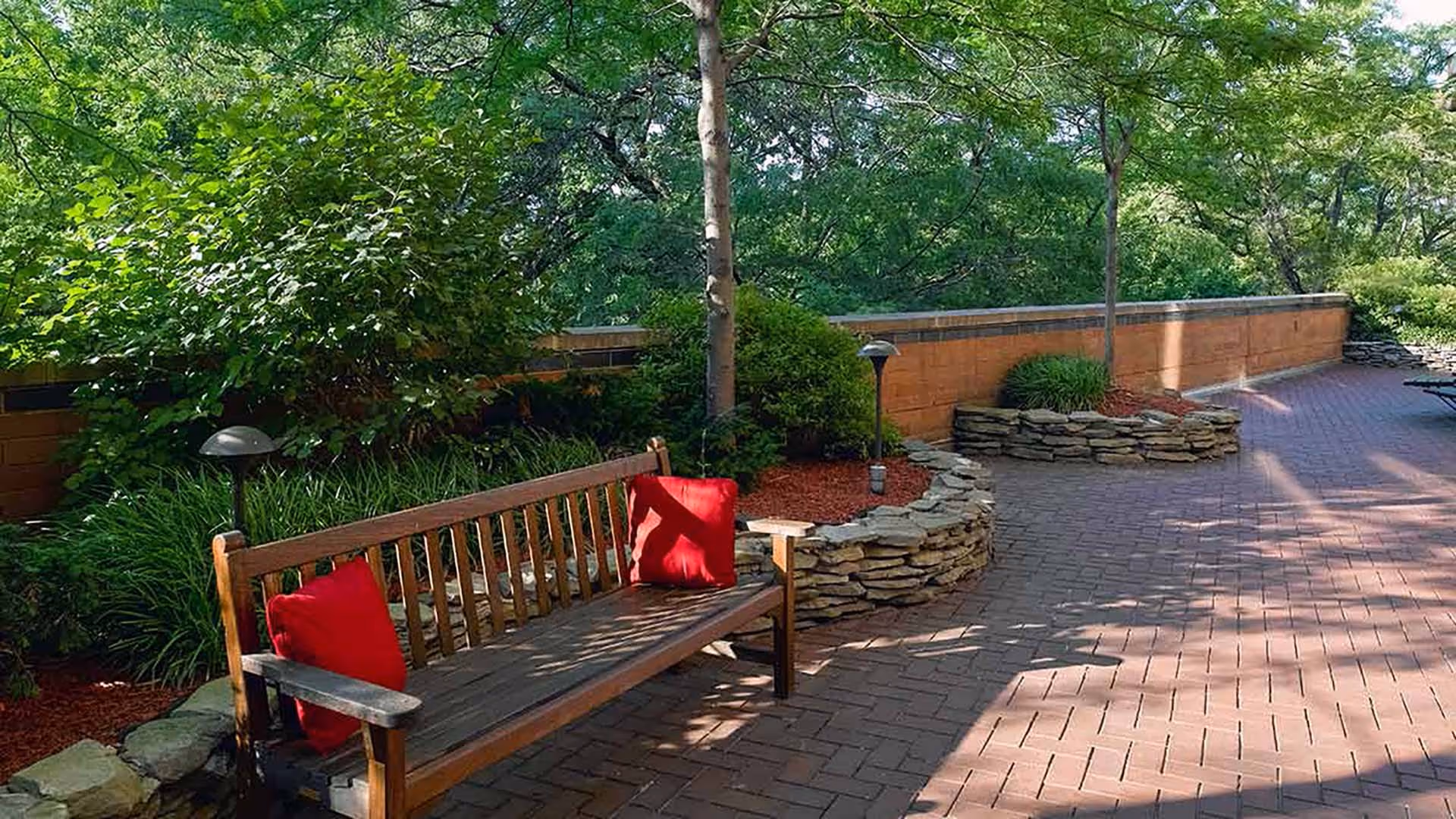 Outdoor patio area with a wooden bench featuring two red cushions, surrounded by green trees and bushes, stone planters, and a brick-paved walkway.