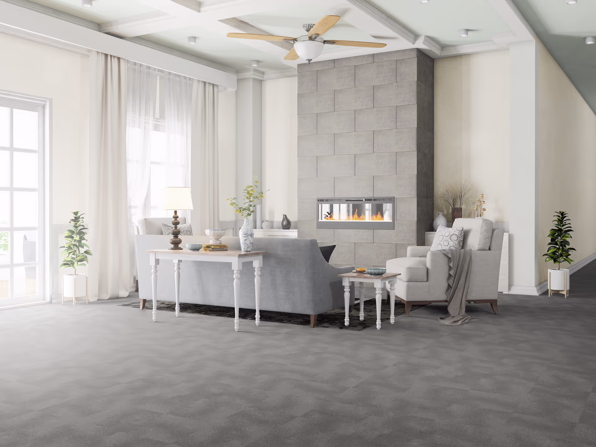 A bright and spacious living room with a modern gray sofa and armchair arranged around a rectangular fireplace set in a tiled wall. The room features large windows with sheer white curtains, a ceiling fan with wooden blades, a gray carpeted floor, and decorative plants and vases placed around the space.