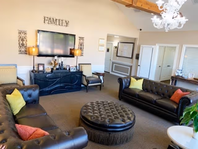 A cozy living room with two dark brown leather tufted sofas adorned with colorful pillows, a large round tufted ottoman in the center, a flat-screen TV mounted on the wall above a black console table, and decorative lamps on either side. The wall above the TV has the word 'FAMILY' displayed, and there is a chandelier hanging from the ceiling.