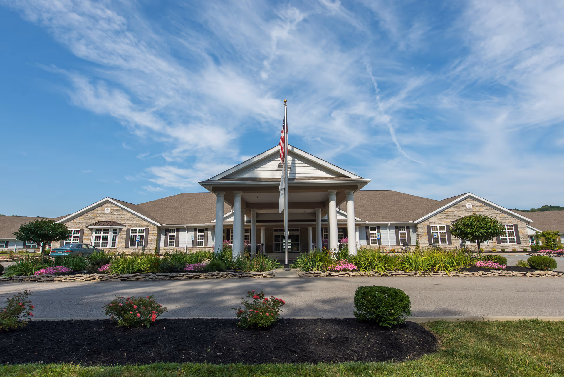 Front exterior view of a single-story senior living facility building with a covered entrance supported by white columns, an American flag on a flagpole in front, landscaped flower beds, and a clear blue sky with wispy clouds above.