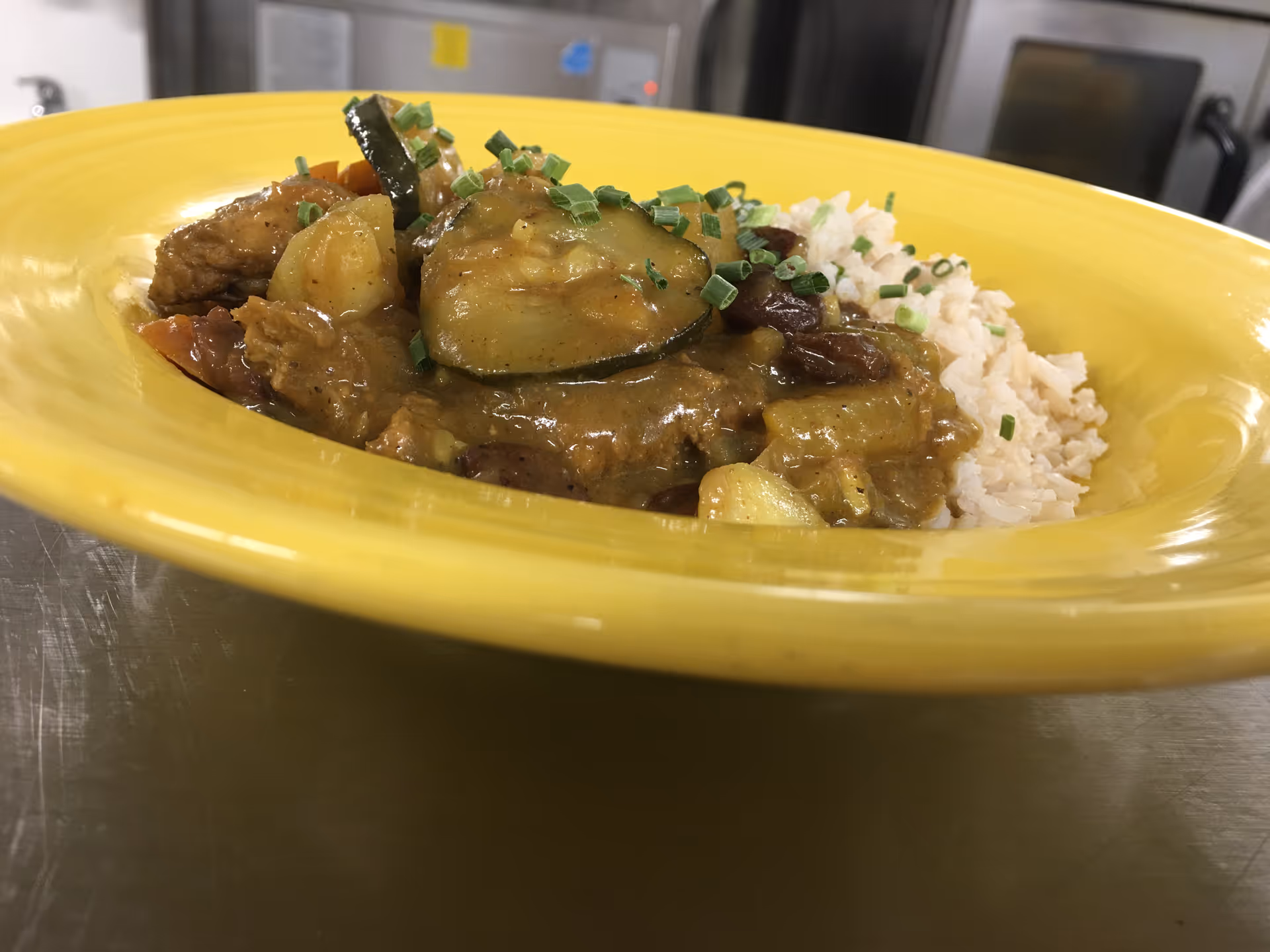 A close-up of a yellow bowl containing a serving of rice topped with a vegetable and meat stew garnished with chopped chives, placed on a stainless steel surface with kitchen equipment in the background.