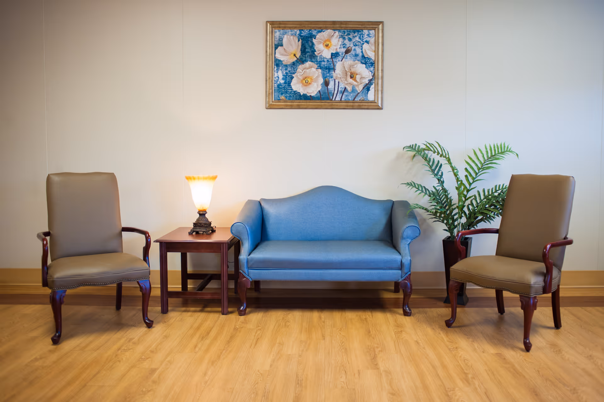 A small waiting area with a blue loveseat flanked by two armchairs, a side table with a lamp, a potted plant, and a floral painting on the wall.