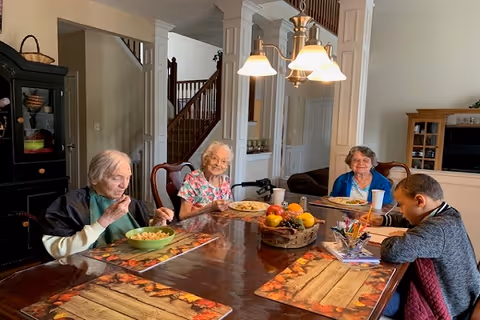 Four elderly individuals sitting around a wooden dining table with autumn-themed placemats. Three of them are eating from bowls and plates, while one person is writing or drawing on paper. The room has a staircase in the background and a cabinet with dishes on the left side.