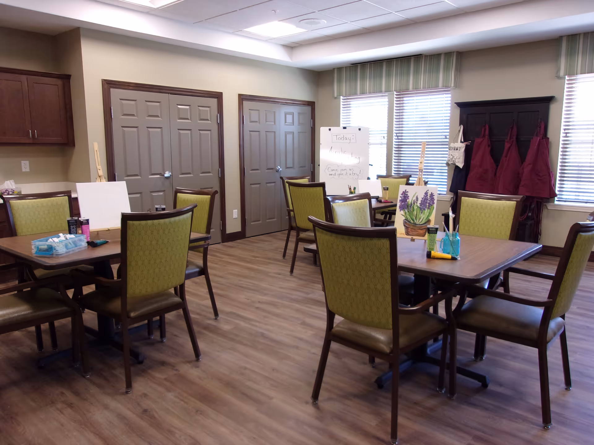 A well-lit activity room with wooden floors and beige walls featuring several tables and green cushioned chairs. On the tables are painting supplies, canvases, and a small easel with a painting of purple flowers. In the background, there is a whiteboard with a message about a morning painting activity and several maroon aprons hanging on hooks near windows with blinds.