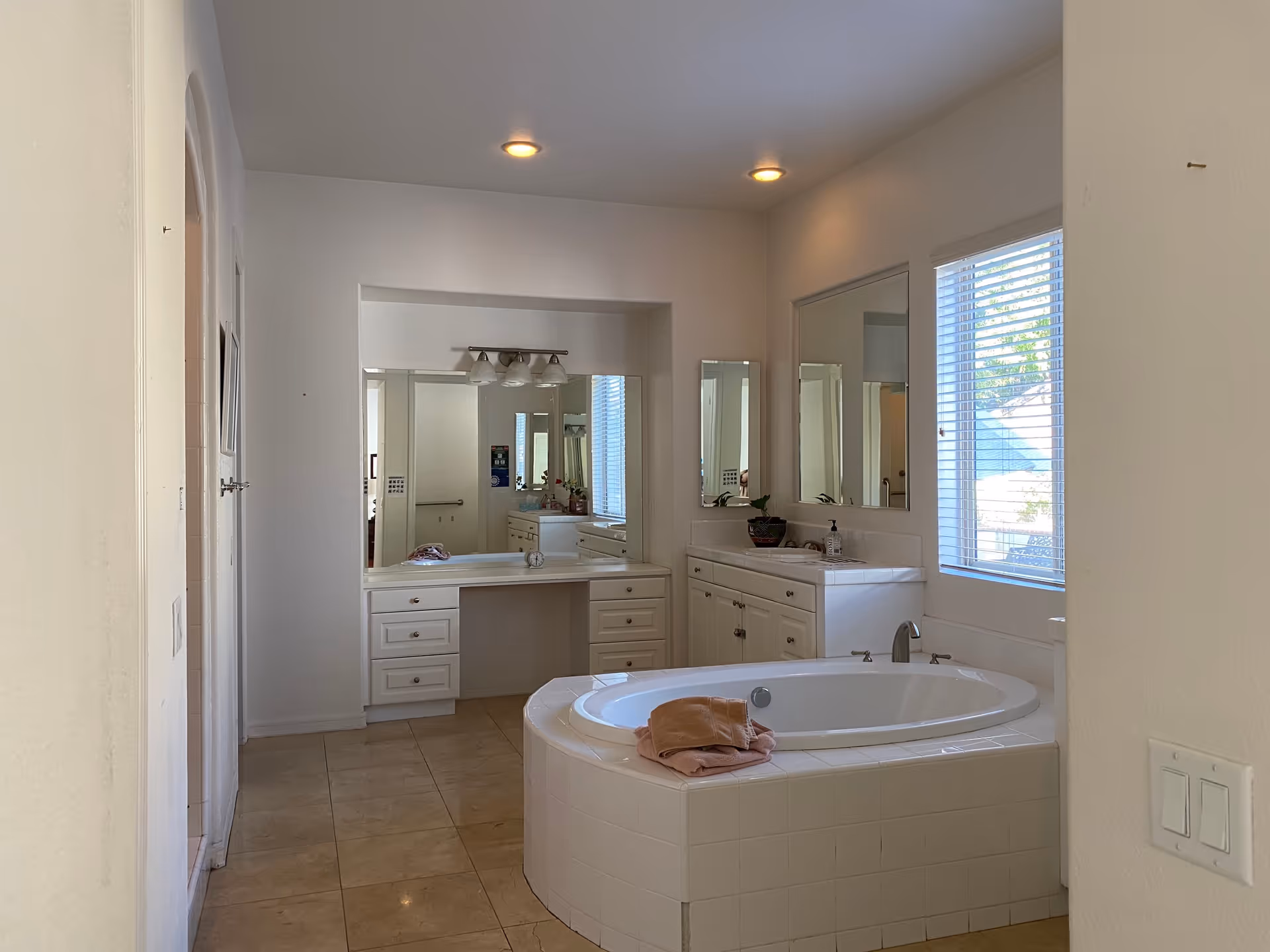 A spacious bathroom with a large white tiled bathtub in the center, a vanity area with multiple mirrors and drawers, and a window with blinds letting in natural light.