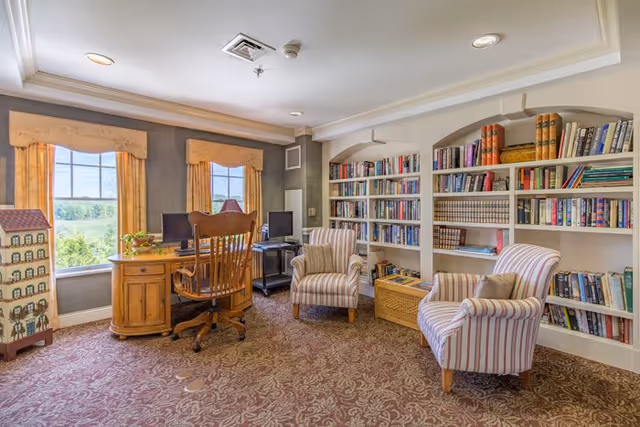 A cozy reading and computer area in a senior living facility with two large windows letting in natural light. The room features a wooden desk with two computer monitors and a wooden chair, two striped upholstered armchairs with cushions, a bookshelf filled with books, and a patterned carpet. The walls are painted gray with white trim and crown molding.