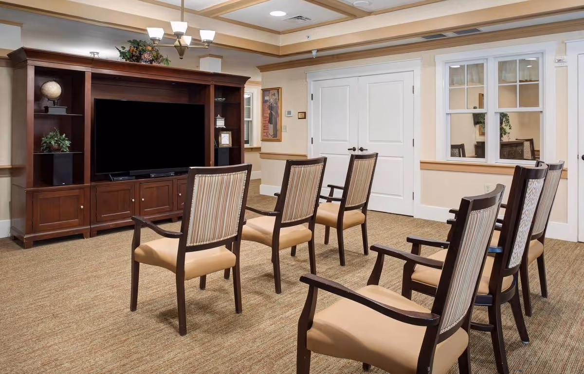 A small meeting or media room with six wooden chairs arranged in two rows facing a large flat-screen TV mounted on a dark wooden entertainment center. The room has beige carpet, cream-colored walls with wooden trim, a double white door, and a window reflecting part of the room.