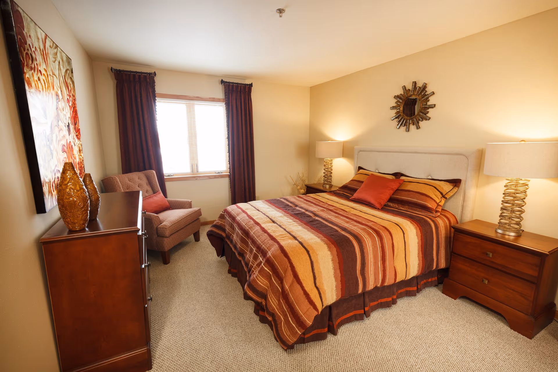 Well-lit bedroom with a bed dressed in striped warm-toned bedding, two nightstands with lamps, a dresser, and an armchair by the window.