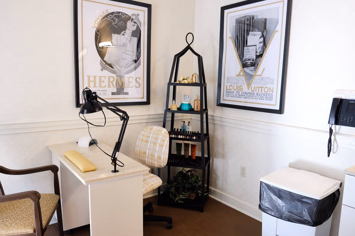 Small salon/nail station with a desk and lamp, shelving of beauty products, two chairs, framed Hermes and Louis Vuitton prints on the wall, and a trash bin.