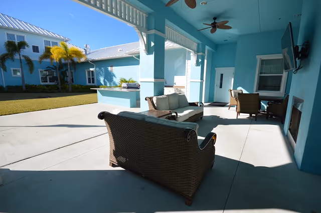 Outdoor covered patio area with wicker furniture including a sofa and chairs, a mounted flat-screen TV, ceiling fans, and a built-in grill. The area is adjacent to a blue building with windows and palm trees visible in the background under a clear blue sky.