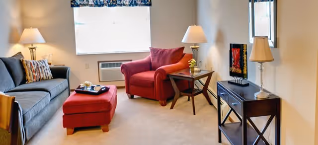 A cozy living room with a gray sofa adorned with patterned pillows, a red armchair with a matching ottoman, and a wooden side table with a lamp and small plant. Another wooden console table with a lamp and decorative items is against the wall, with a window covered by a valance letting in natural light.