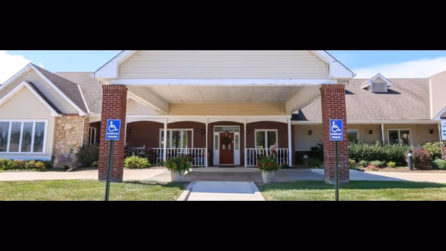 Front exterior view of Vintage Park at Baldwin City building with a covered entrance supported by brick columns, two handicap parking signs, and landscaped greenery around the entrance.