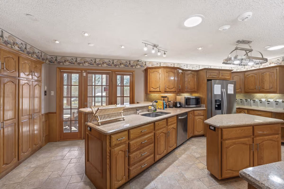 A spacious kitchen with wooden cabinets and drawers, a central island, and a countertop with a dish rack. The kitchen features stainless steel appliances including a refrigerator, dishwasher, and microwave. There are French doors leading outside, tile flooring, and decorative wallpaper border near the ceiling.