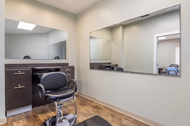 Interior view of a small salon or grooming area with a black salon chair in front of a dark brown vanity with drawers and a large mirror above it. Another large mirror is mounted on the adjacent wall, reflecting the room and part of a doorway leading to another room with chairs.