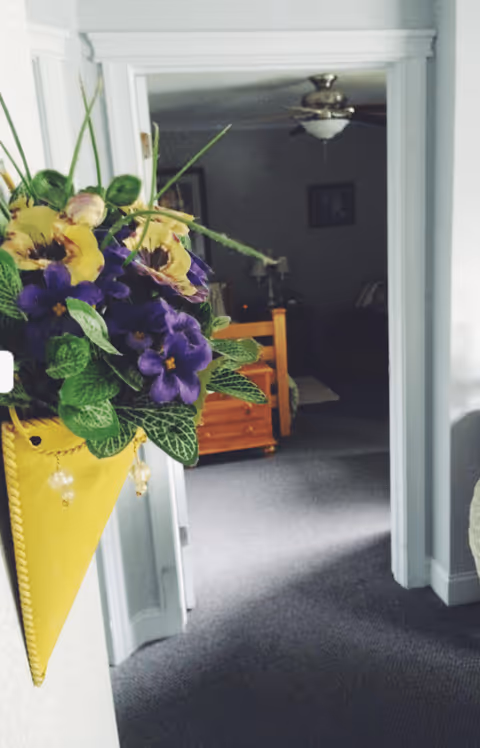 View down a hallway with a yellow wall-mounted vase holding purple and yellow flowers on the left. The hallway leads to a dimly lit room with a wooden bed frame, a ceiling fan, and framed pictures on the wall.