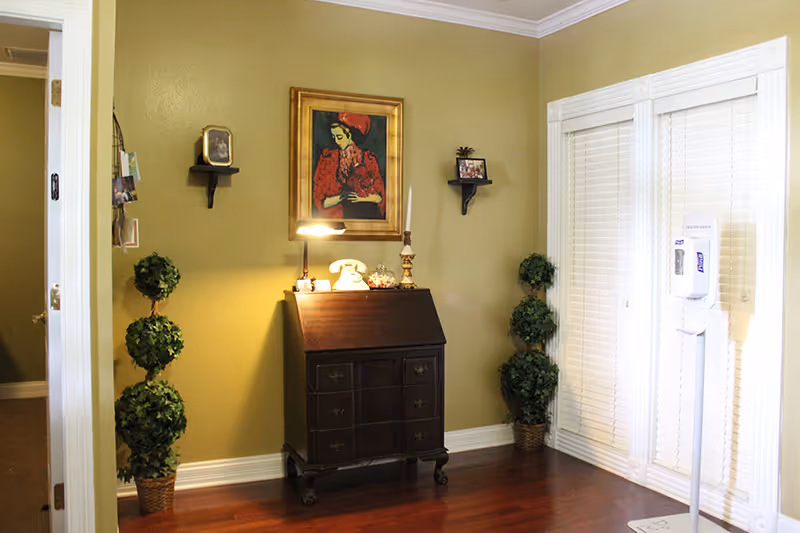 A corner of a room with olive green walls and wooden flooring. There is a dark wooden cabinet with drawers and a slanted top, on which sits a vintage white rotary phone, a lamp, and decorative items. Above the cabinet hangs a framed painting of a woman in a red dress. On either side of the cabinet are two potted topiary plants. Two small wall shelves hold framed photos. To the right, there are white blinds covering a window or door, and a hand sanitizer dispenser stands in front of them.