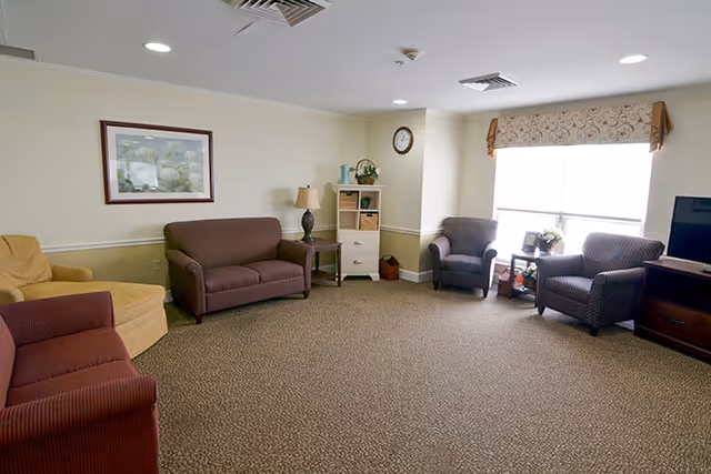 Sunny communal living room with sofas, armchairs, side tables, a TV, and a window letting in natural light.