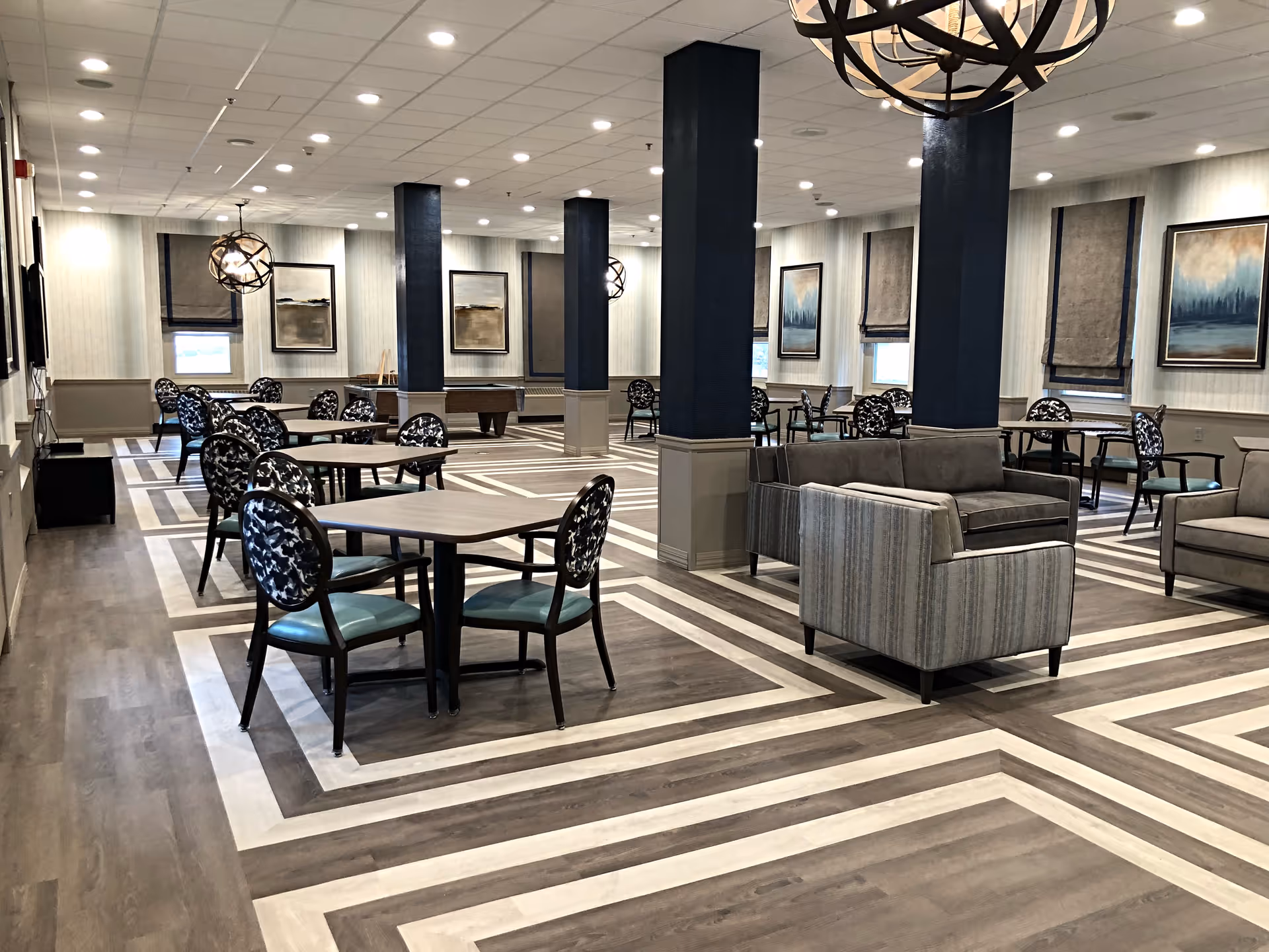 A spacious, well-lit common area with multiple tables and chairs arranged for seating, along with comfortable sofas. The room features patterned flooring, several dark blue pillars, modern light fixtures, and framed artwork on the walls with windows covered by Roman shades.