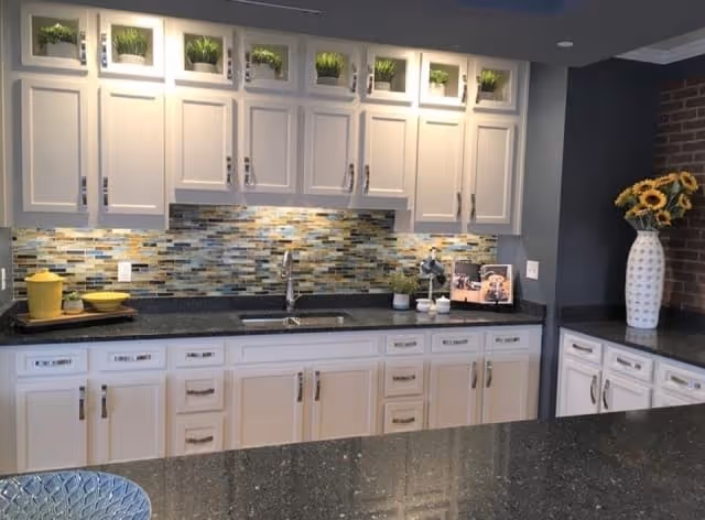Modern kitchen with white cabinets, a mosaic tile backsplash in shades of blue, gray, and beige, a stainless steel sink with a faucet, and a dark granite countertop. There are decorative plants above the upper cabinets and a vase with sunflowers on the right side countertop.