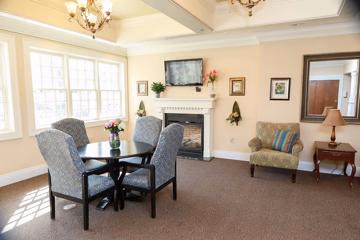 Sunny senior living common room with a round table and four chairs, a fireplace with a TV above, and an upholstered armchair.