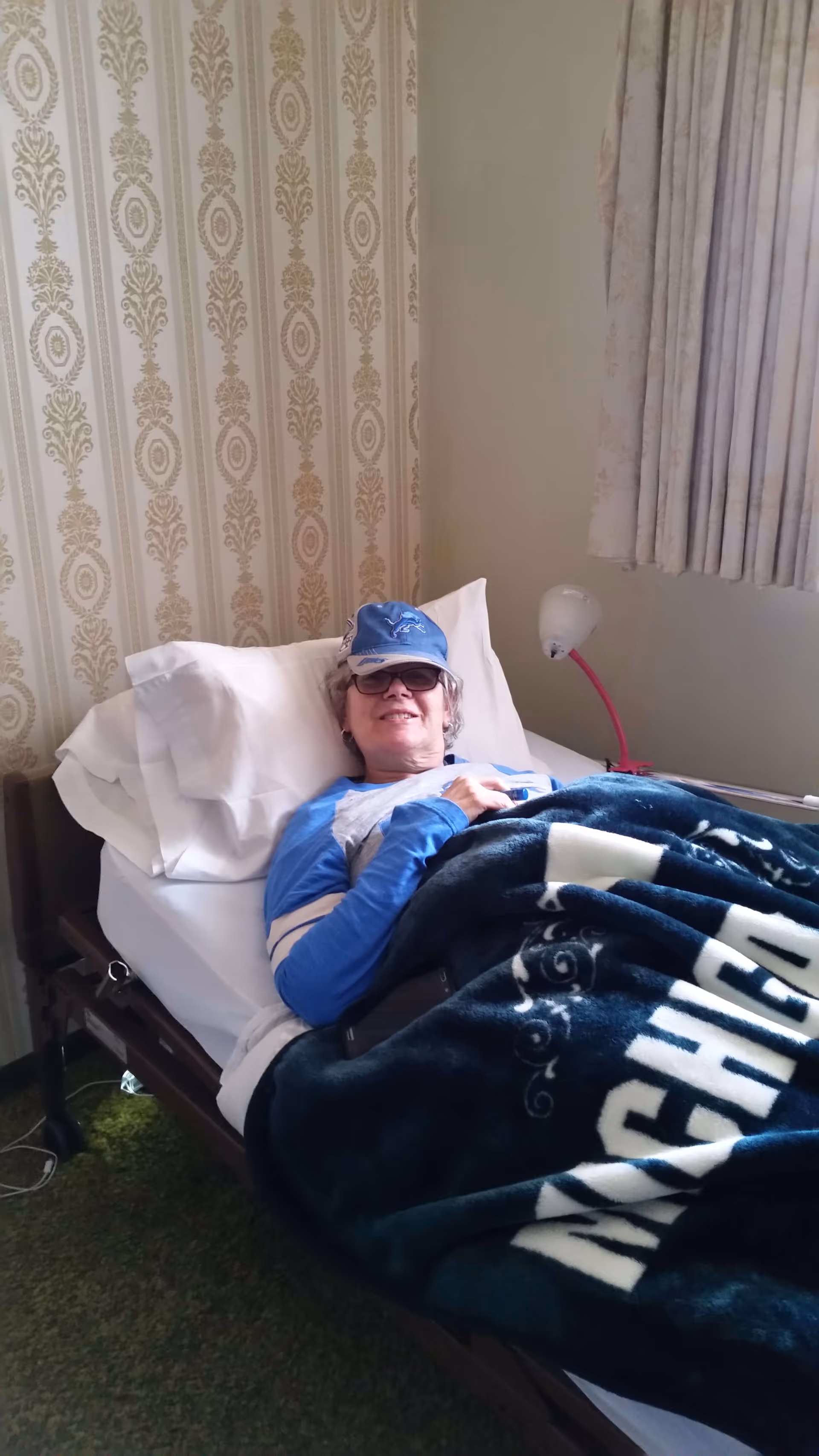 An elderly person lying in a bed covered with a dark blue blanket that has white text. The person is wearing glasses, a blue long-sleeve shirt, and a blue cap. The bed is positioned against a wall with patterned wallpaper and next to a window with light-colored curtains. A small lamp with a red flexible neck is attached to the wall near the bed.