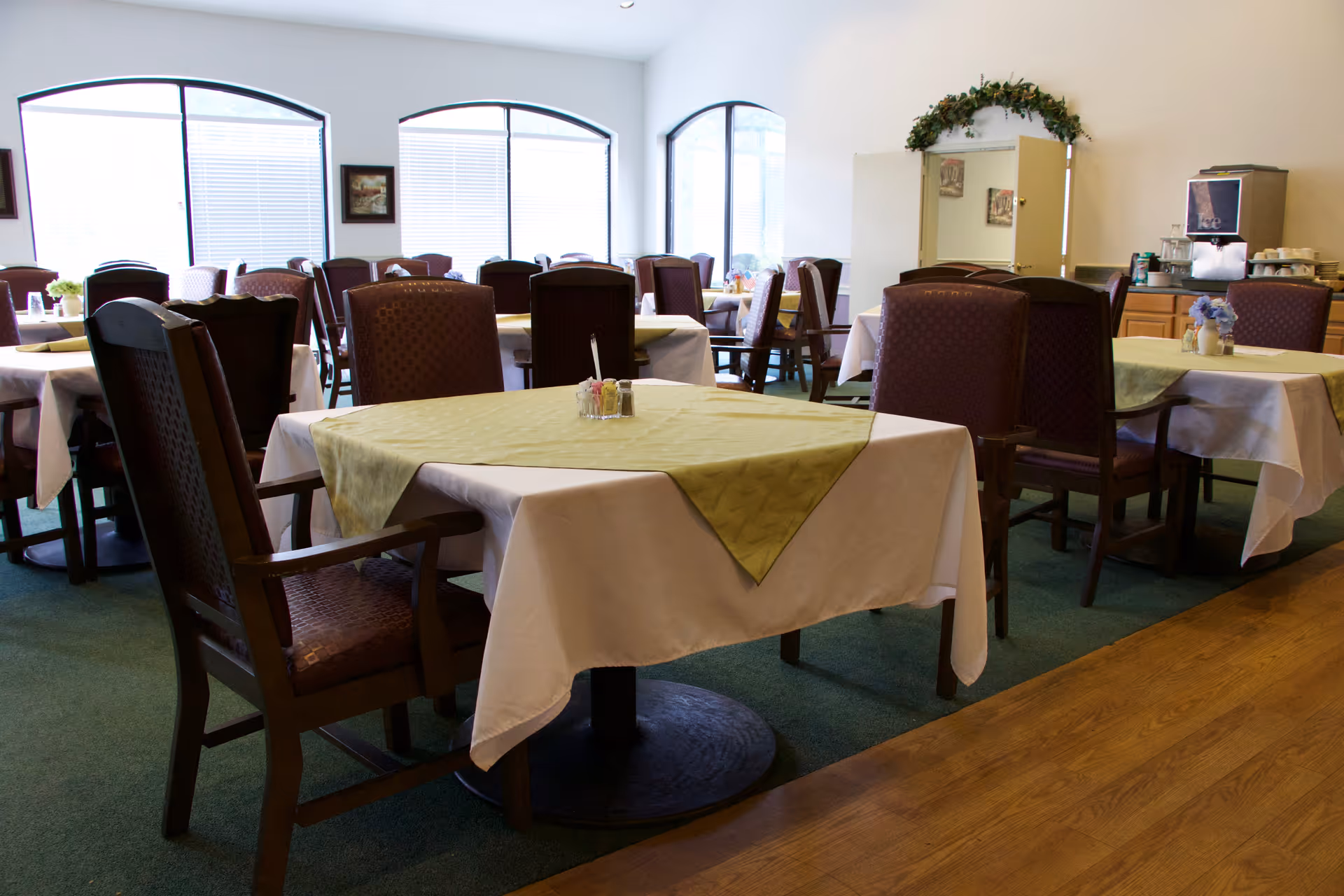 A dining room with multiple tables covered with white tablecloths and green table runners. Each table is surrounded by wooden chairs with cushioned seats and backs. The room has large arched windows letting in natural light, and there is a beverage station with a coffee machine and cups in the background.