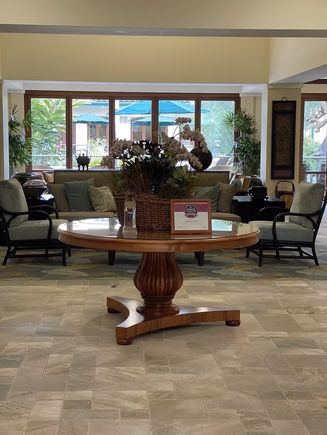 Lobby interior with a round wooden table centerpiece holding orchids and a plaque, sofas and armchairs in front of large windows showing patio umbrellas.