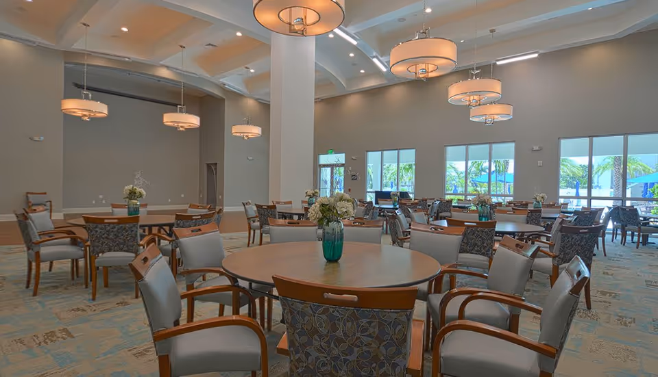 A spacious dining room with multiple round wooden tables surrounded by cushioned chairs. Each table has a vase with flowers as a centerpiece. Large windows allow natural light to fill the room, and modern pendant lights hang from the high ceiling.