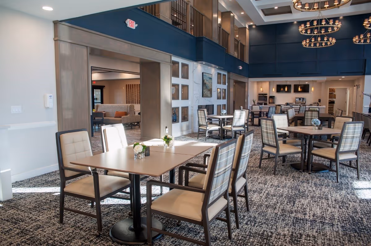 A spacious and well-lit dining area with multiple tables and chairs arranged neatly. The room features a high ceiling with modern chandeliers, a carpeted floor, and a kitchen area in the background with appliances and countertops. There is a seating area visible through a large open doorway, and decorative elements such as flowers on the tables and framed artwork on the walls.