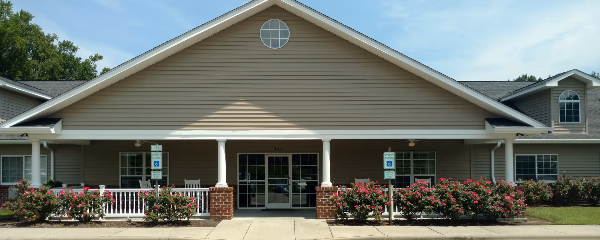 Front entrance of a single-story senior living building with a covered porch, white columns, double glass doors, and flowering shrubs.