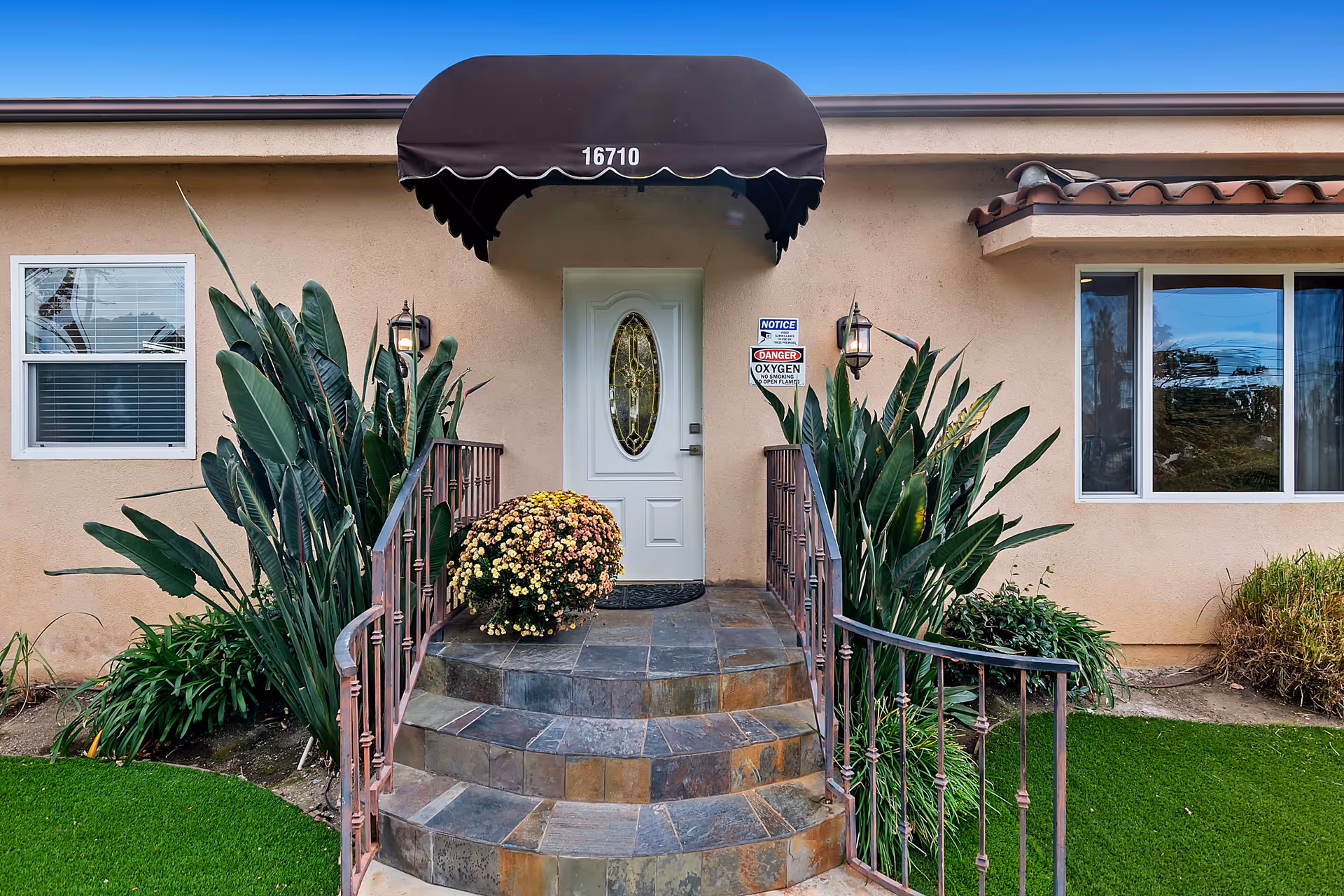Front entrance of a single-story building with tiled steps, a brown awning labeled "16710", a white oval-paneled door, potted flowers and surrounding plants.