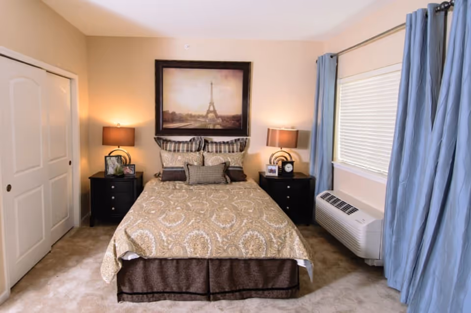 Sunlit bedroom with a centered bed dressed in patterned linens, matching nightstands with lamps, a framed Eiffel Tower picture above, and a window with blue curtains.