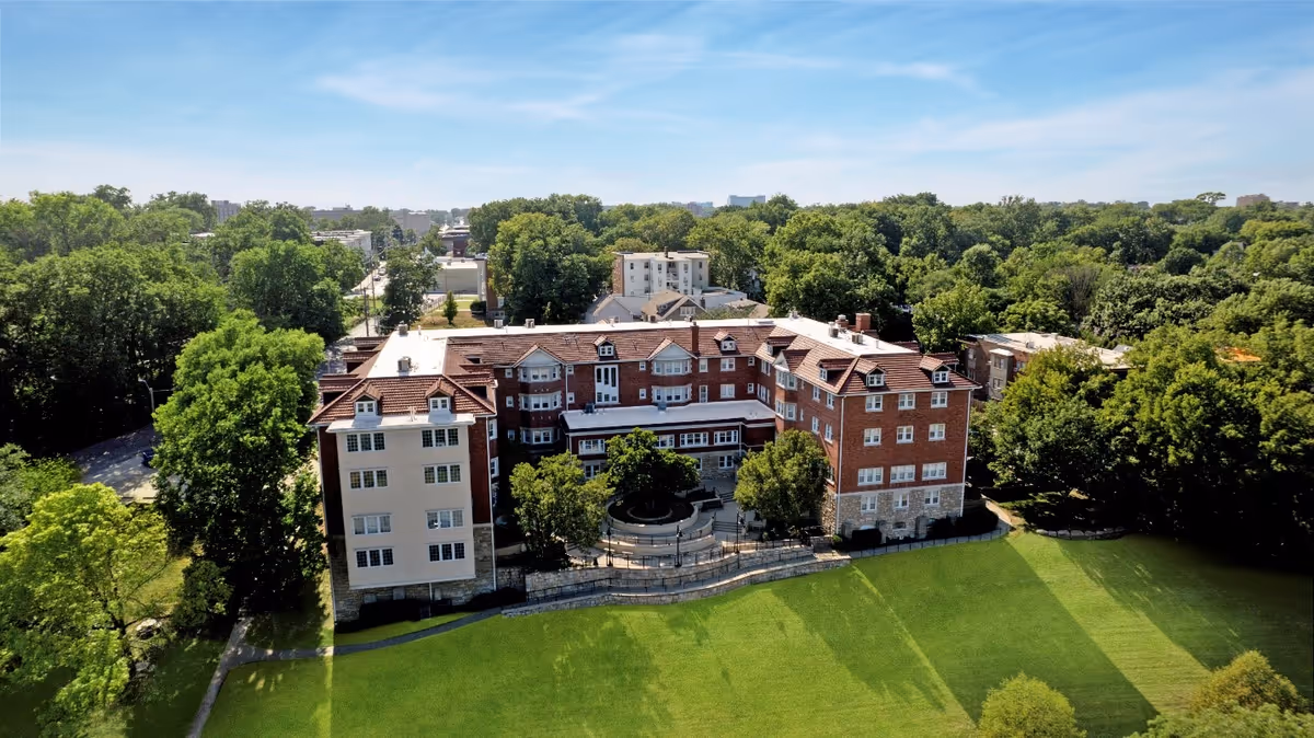 Aerial view of Rockhill Manor, a large multi-story brick building with a red-tiled roof, surrounded by green trees and a well-maintained lawn under a clear blue sky.