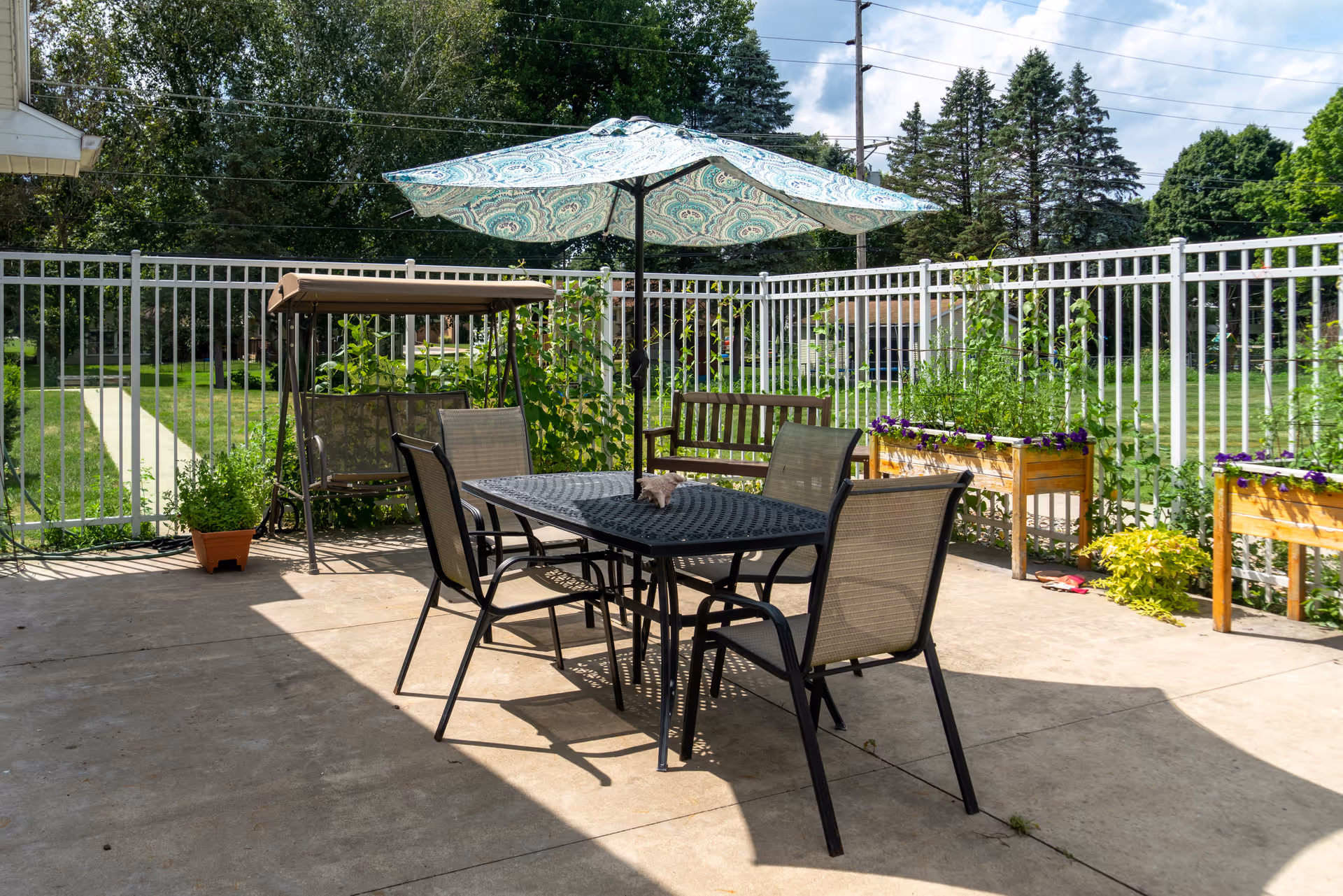Outdoor patio area with a black metal table and four chairs, a large patterned umbrella providing shade, a wooden bench, a swing with a canopy, and several planter boxes with flowers and greenery, all enclosed by a white metal fence with trees and grass visible beyond.