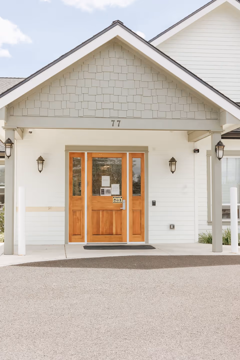 Front entrance of a building with a wooden double door, white exterior walls, and a gabled roof. The number 77 is displayed above the door, and there are wall-mounted lantern-style lights on either side of the entrance.