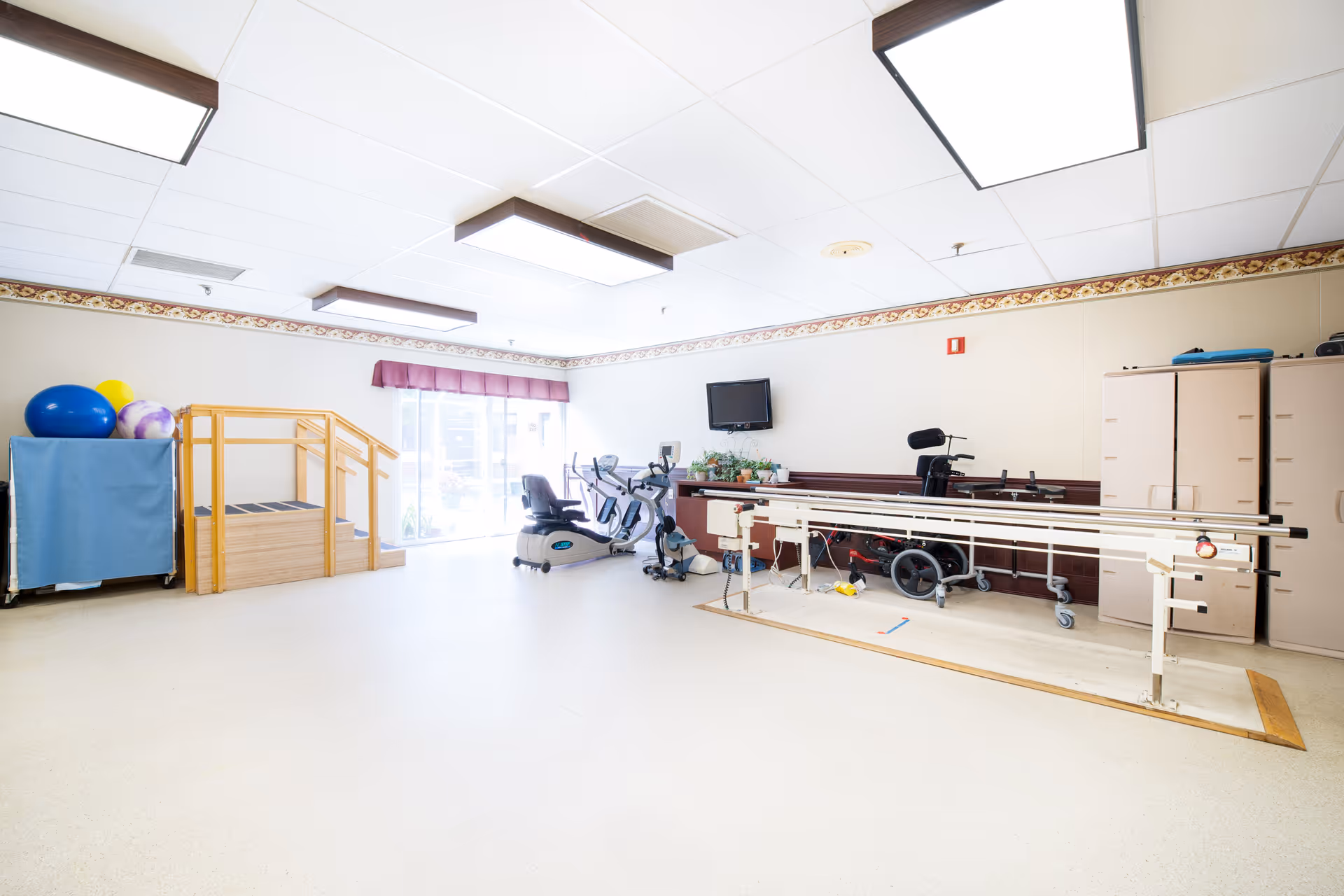 A bright therapy room with exercise equipment including stationary bikes, parallel bars for walking practice, a wheelchair, therapy balls on a rack, and a small wooden staircase. The room has a large window with a purple valance, beige walls with a decorative border near the ceiling, and overhead fluorescent lighting.
