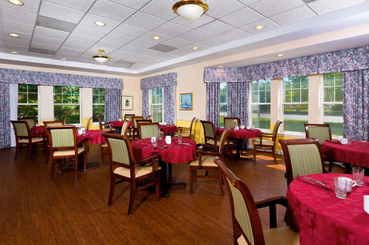 A dining room with multiple round tables covered with red tablecloths, each set with cups, glasses, and utensils. The room has wooden chairs with green striped cushions, large windows with floral curtains, and a wooden floor. The ceiling has recessed lighting and two ceiling light fixtures.