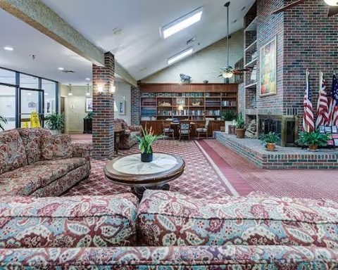 Community lounge with patterned sofas, a round coffee table, brick fireplace, and a bookcase under a vaulted ceiling.