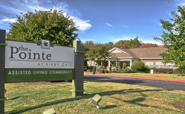 Outdoor view of The Pointe at Kirby Gate Assisted Living Community sign with a single-story building and trees in the background under a blue sky with some clouds.