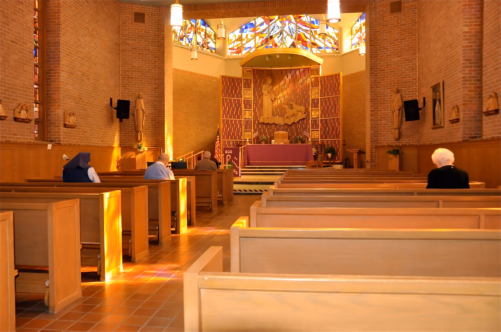Interior of a chapel with wooden pews and a few elderly people seated. The chapel has brick walls, stained glass windows near the ceiling, religious statues on the walls, and an altar at the front with religious decorations and an American flag.