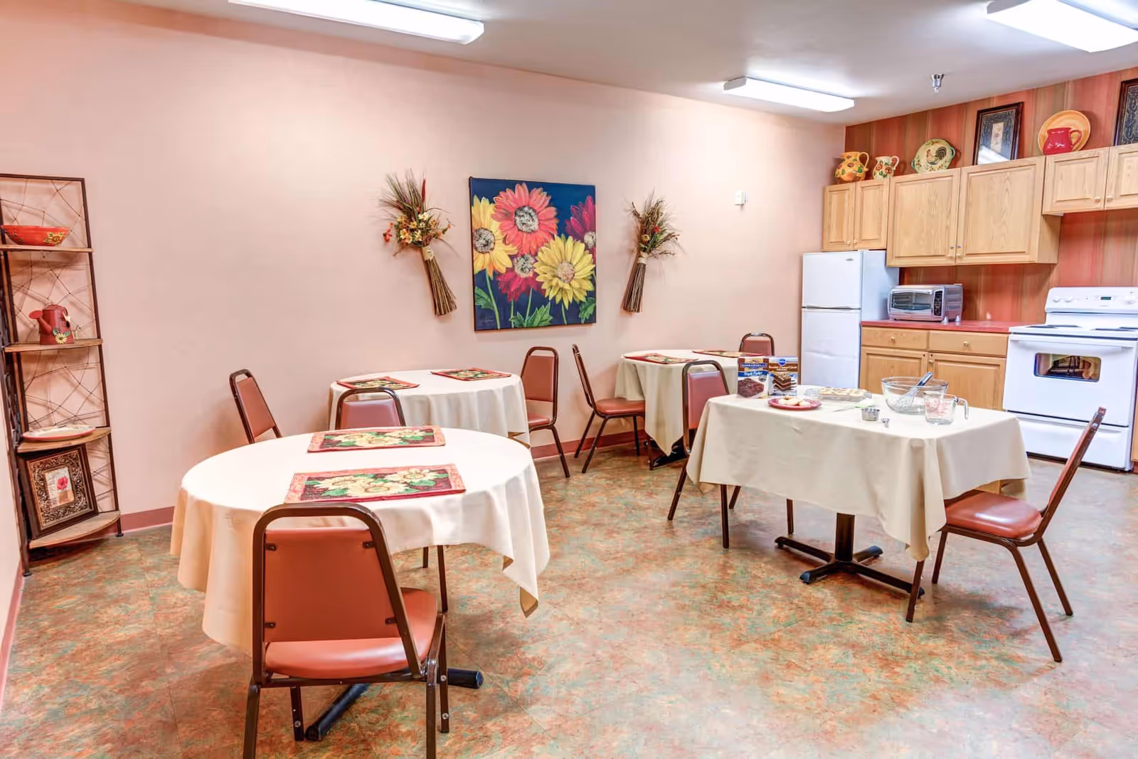 A small dining area with four tables covered in beige tablecloths and floral placemats. The room has pink walls decorated with a colorful flower painting and two floral wall hangings. There are brown chairs around the tables. In the background, there is a kitchen area with wooden cabinets, a white refrigerator, a white stove, a toaster oven, and decorative pottery on top of the cabinets. A shelving unit with decorative items is on the left side of the room.
