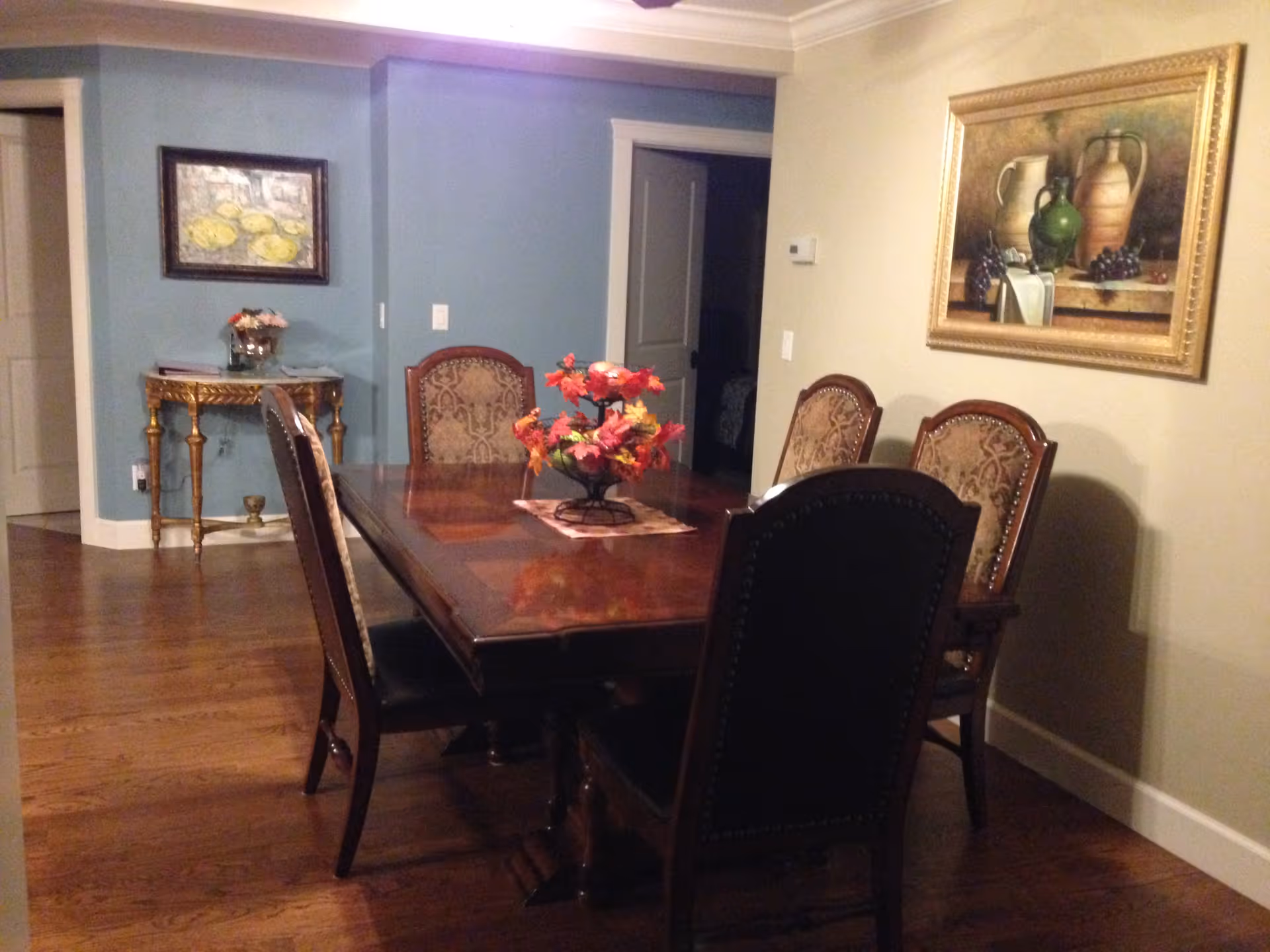 A dining room with a polished wooden table surrounded by six upholstered chairs. A floral centerpiece decorates the table. The room has wooden flooring, a blue accent wall with a small ornate table and a framed painting above it, and another framed painting on the adjacent beige wall.