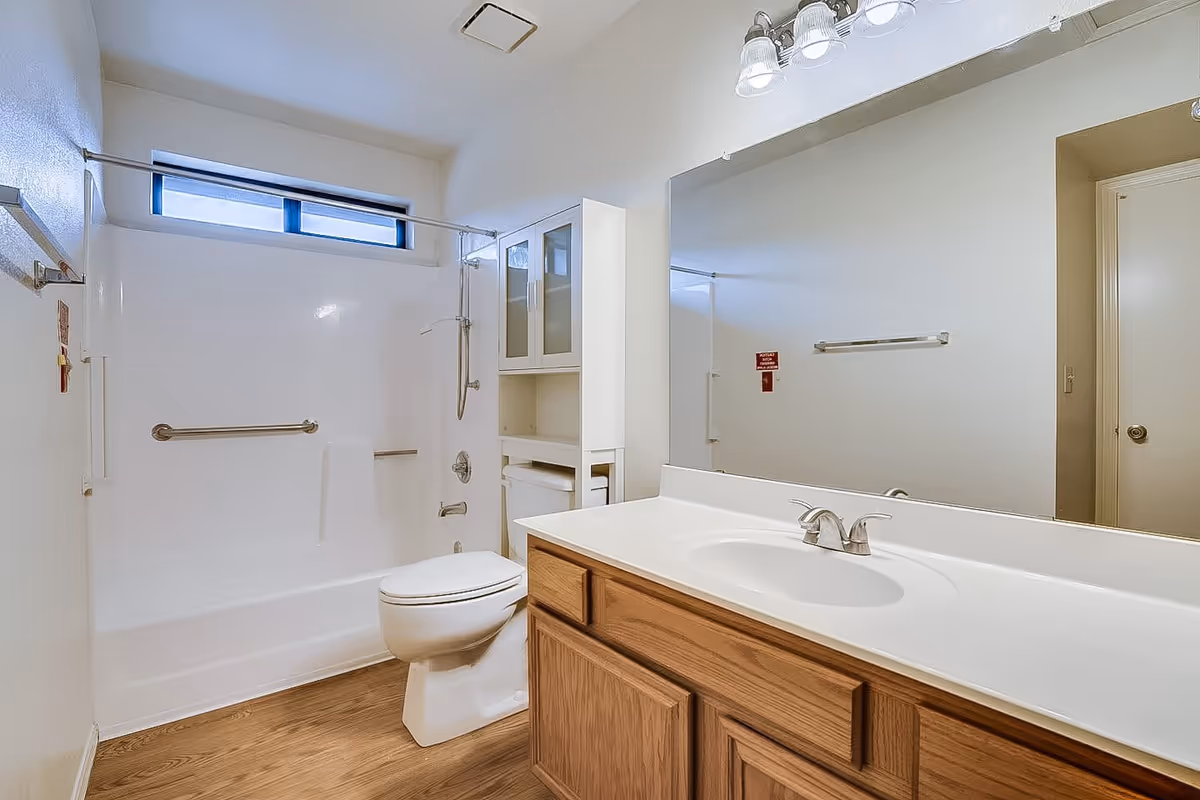 A clean bathroom with a white bathtub and shower combination, a toilet, a wooden vanity with a white countertop and sink, a large mirror above the sink, and a white cabinet with glass doors next to the bathtub. The floor has a wood-like finish and there is a small horizontal window above the bathtub.